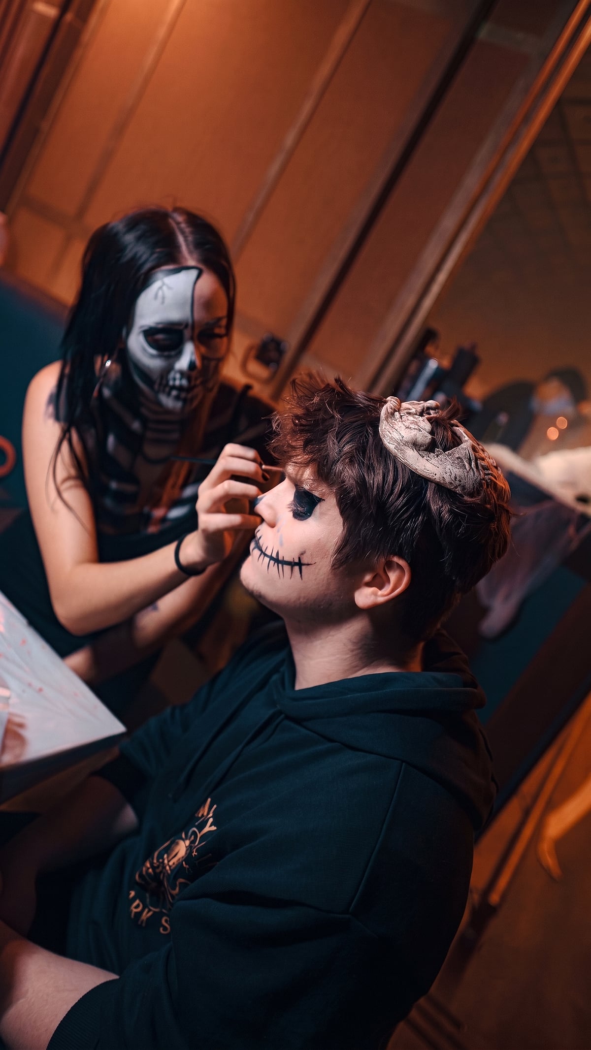 Gaia, in skeleton face paint, painting a stitched-mouth design onto a client at a Halloween night