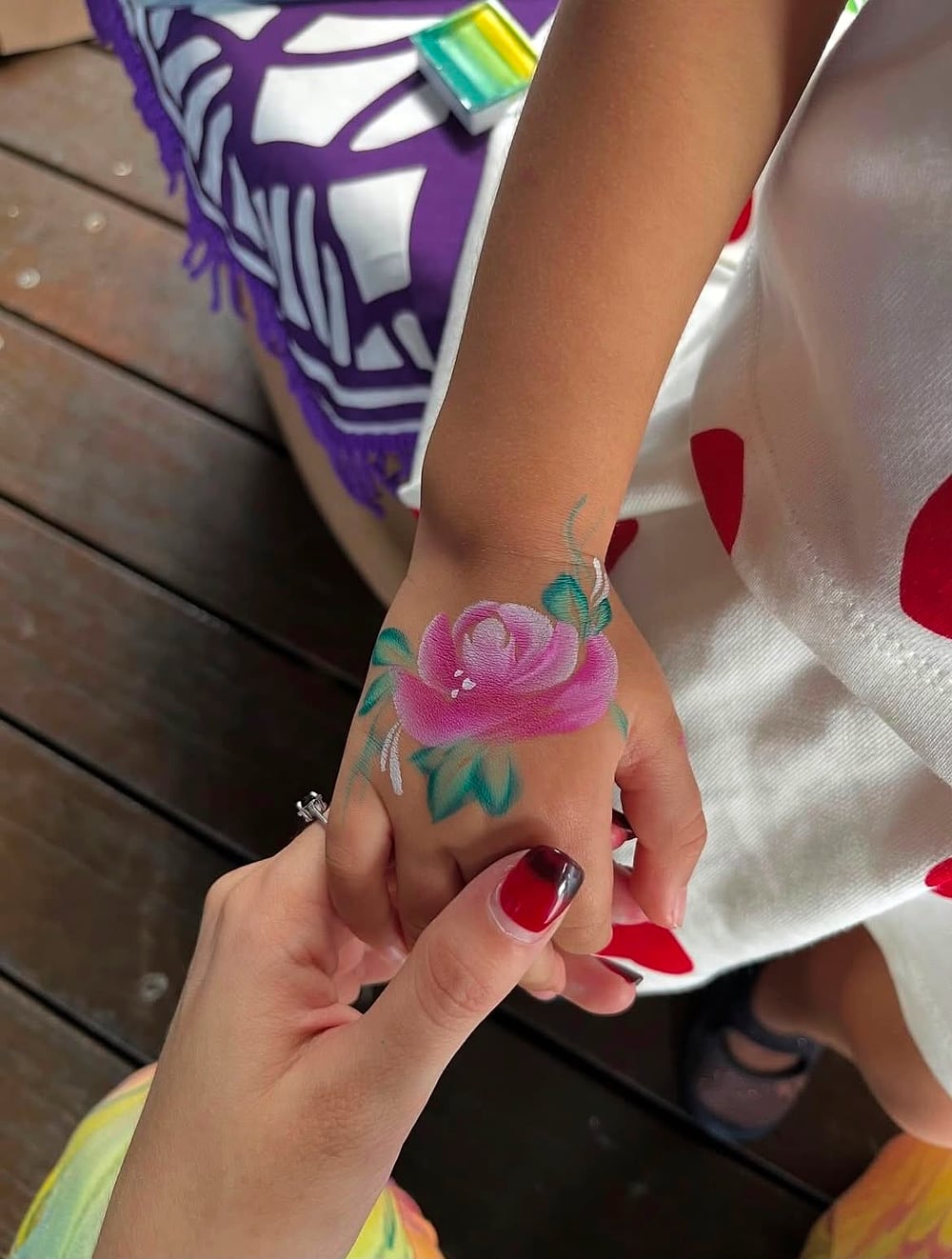 A pink rose painted on the back of a child's hand, held gently by Gaia