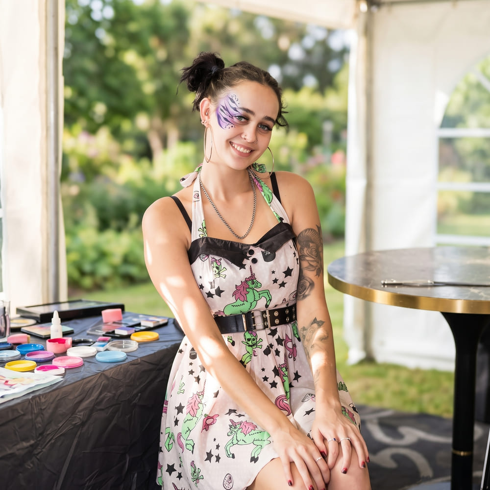 Gaia sitting at her face-painting table under a marquee, paints laid out in front of her